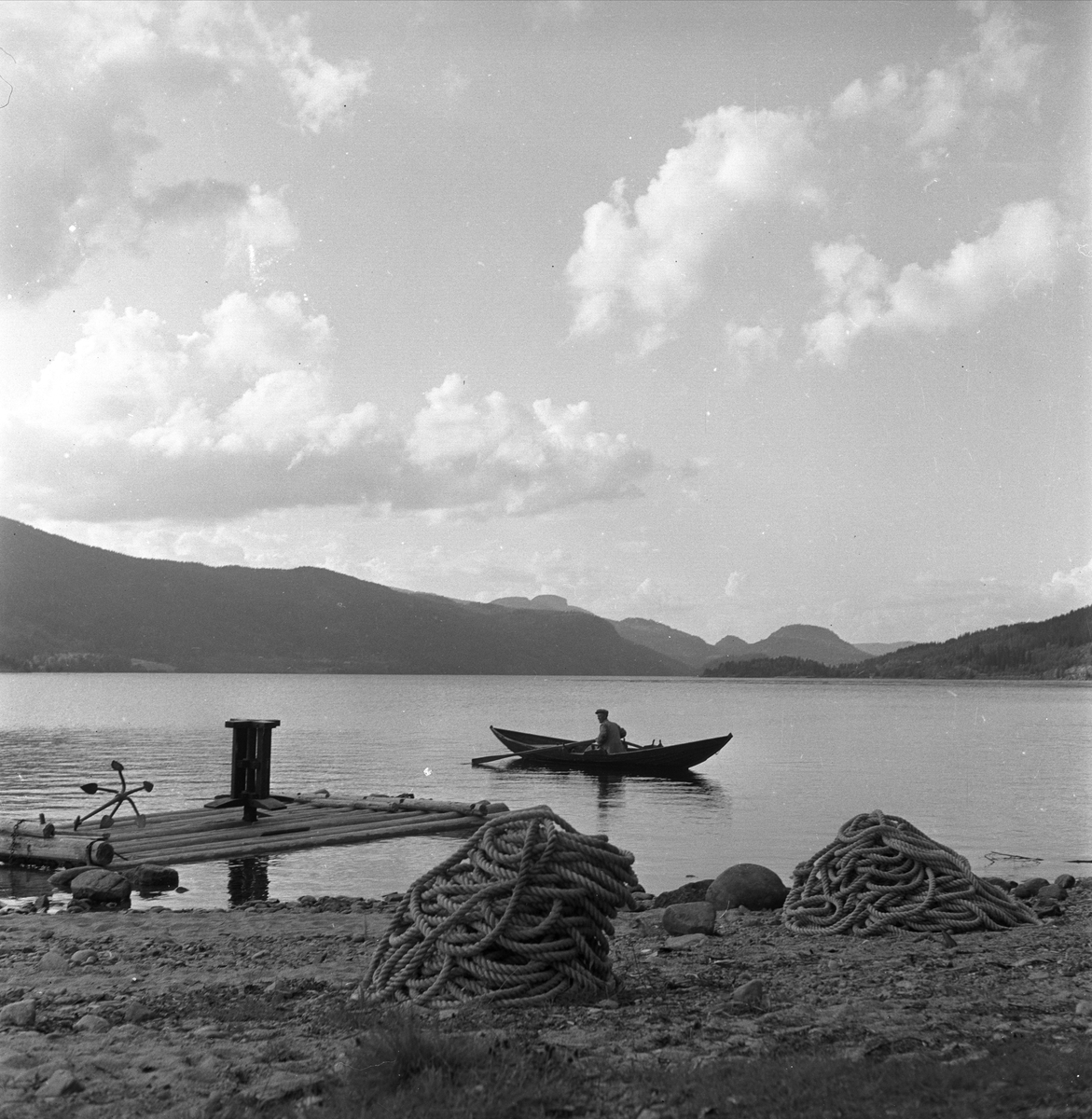 Tømmerfløtere i Sperillen, Ringerike, Buskerud, ant. 1950-tallet. Mann i robåt. Flåte for sleping (bomming) av tømmer.