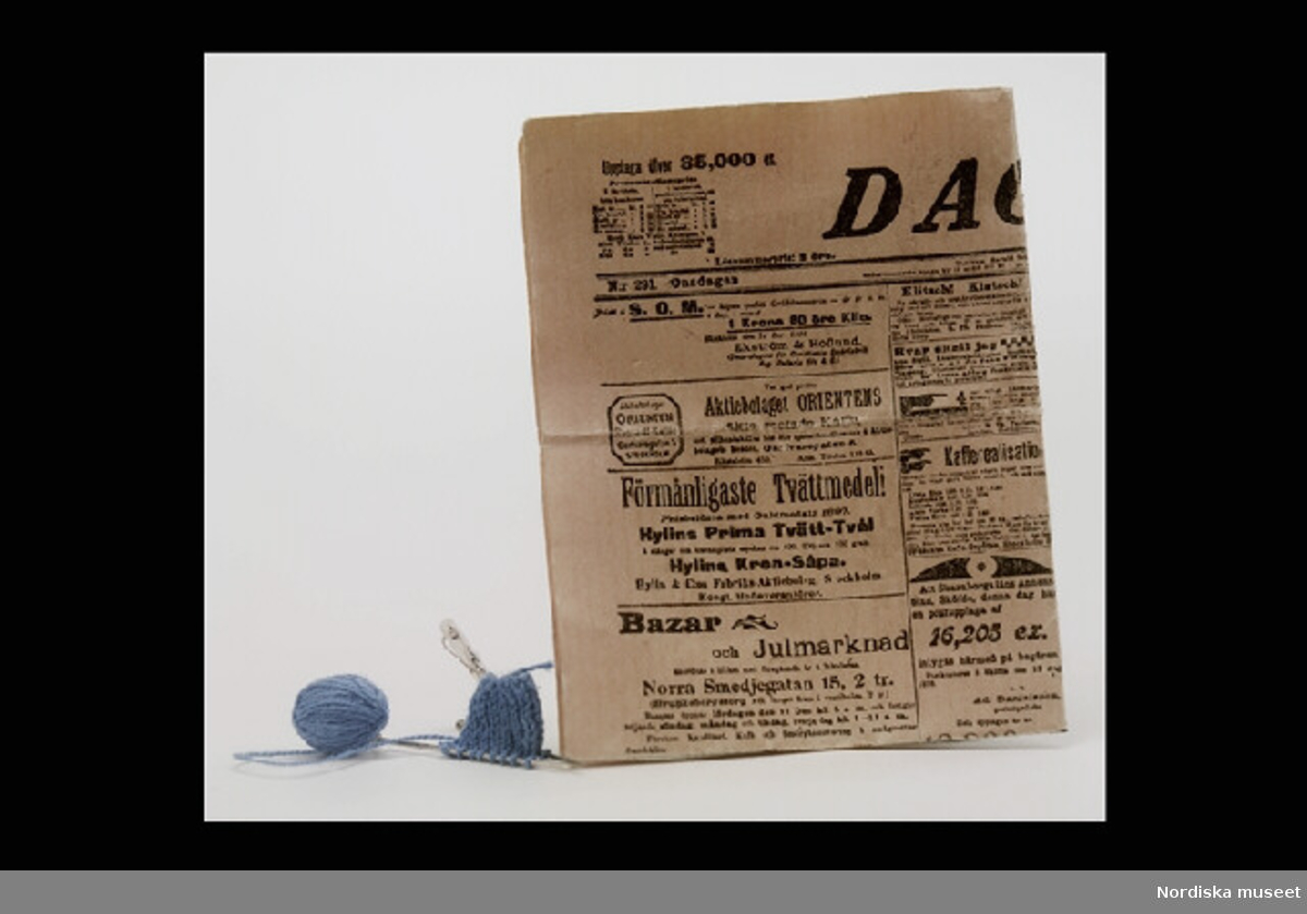 Inventering Sesam 1996-1999:
a) Tidning H 14,6 cm, b 11,3 cm
b) Stickning L (stickor) 2,6 cm
Tidning och stickning.
a) Tidning, tryckt på rosafärgat papper, föreställande framsidan på tidningen "DAGEN" från 14 dec 1898. Troligen extakt kopia på tidningens framsida nämnda dag. En mängd olika annonser och artiklar går att läsa.
b) Stickning av blå tråd med nystan, stickat på två knappnålar. 
Hittad omärkt i dockskåpet (inv.nr 200.513) vid inventering 1997.
Tillhör dockskåp inv.nr 200.513, se dess kat.kort för mer info.
Leif Wallin okt 1997