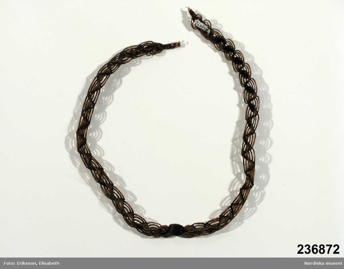 Kedja i glest hårarbete med överklädd kula på mitten,  är ej monterat med metallbeslag och funktionen framgår inte, möjligen halsband eller klockkedja.
/Berit Eldvik 2009-12-01
