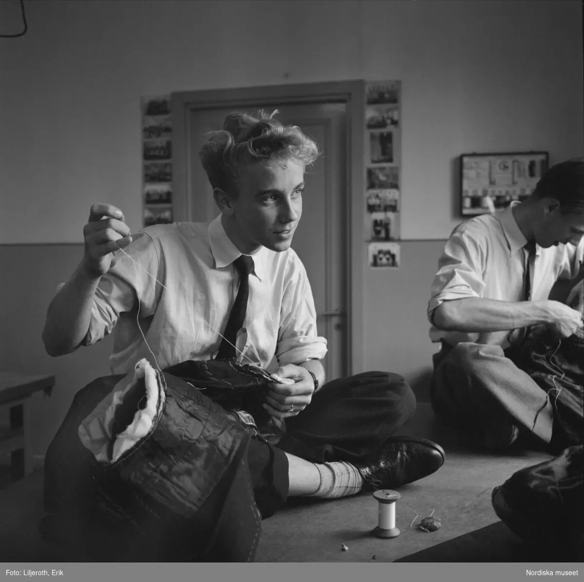 Två unga män sitter och syr på ett bord med benen i kors i "skräddarställning". Borås verkstadsskola för skrädderiyrket.