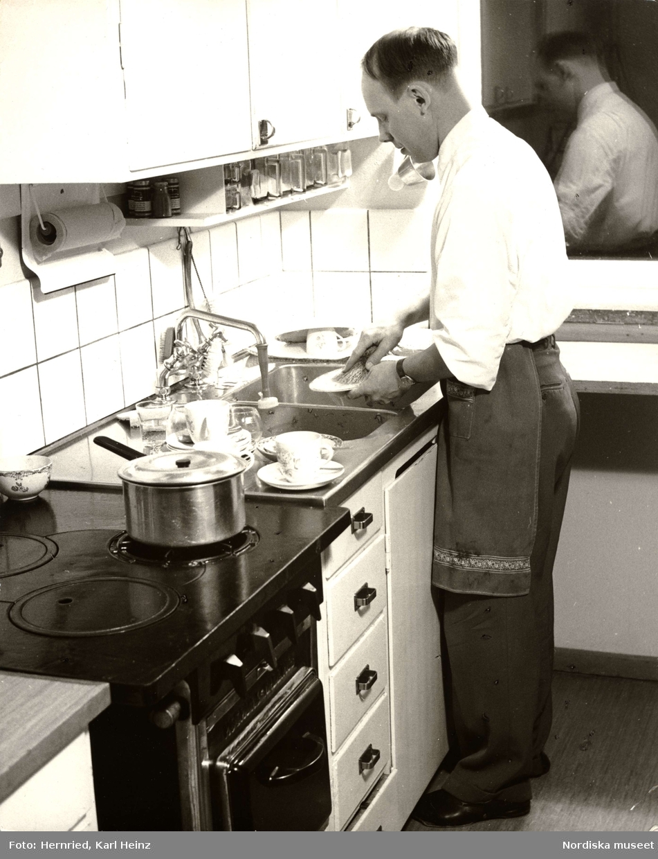 Kök. Interiör.  En man med förkläde står vid diskbänk och diskar. 