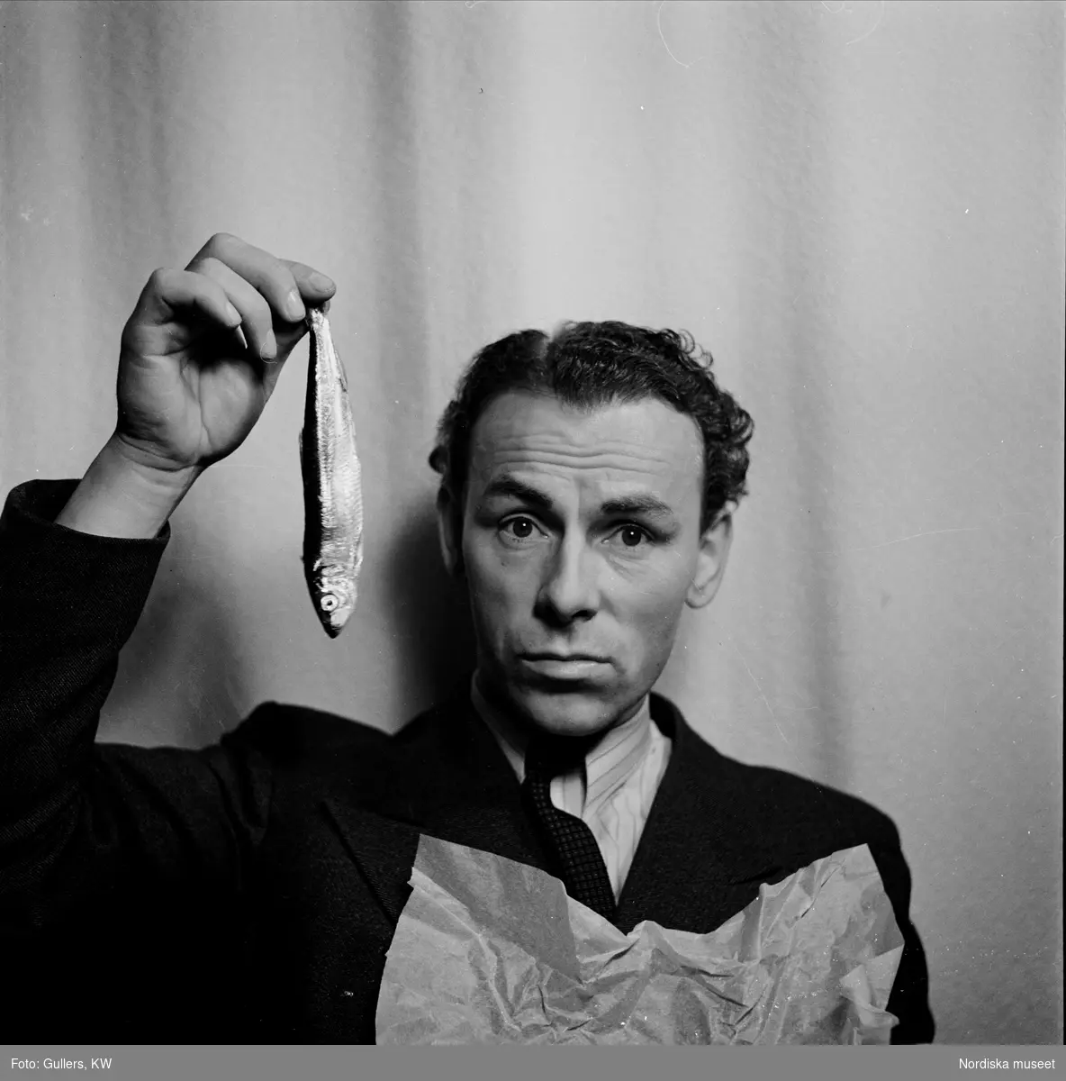 Nils Poppe (1908-2000), skådespelare och komiker, håller upp strömming med handen. - Nordiska museet /