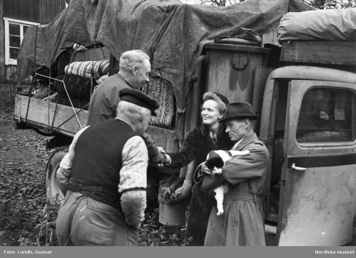 Statare. Paret Eriksson säger adjö till sina grannar vid en flytt. De står framför en lastbi, mannen håller en katt i famnenl.