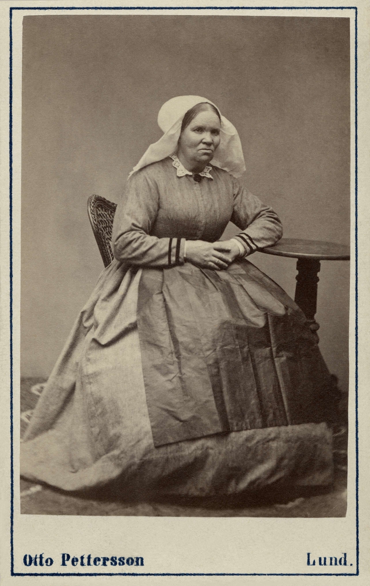 Kvinna poserar i en sockendräkt, sittande med armen vilande på litet pelarbord.  Från Saxtorp, Harjagers härad i Skåne. Inv.nr. 18518N.