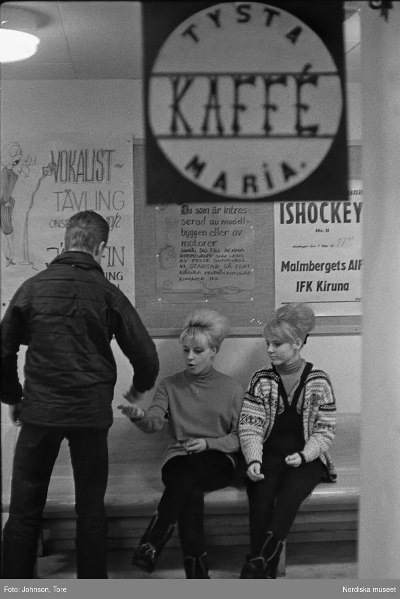Ungdomsgård i Malmberget. Två flickor sitter på bänk, pojke med ryggen mot kameran står framför dem. Anslagstavla med skylt "Tysta  Maria Kaffé".