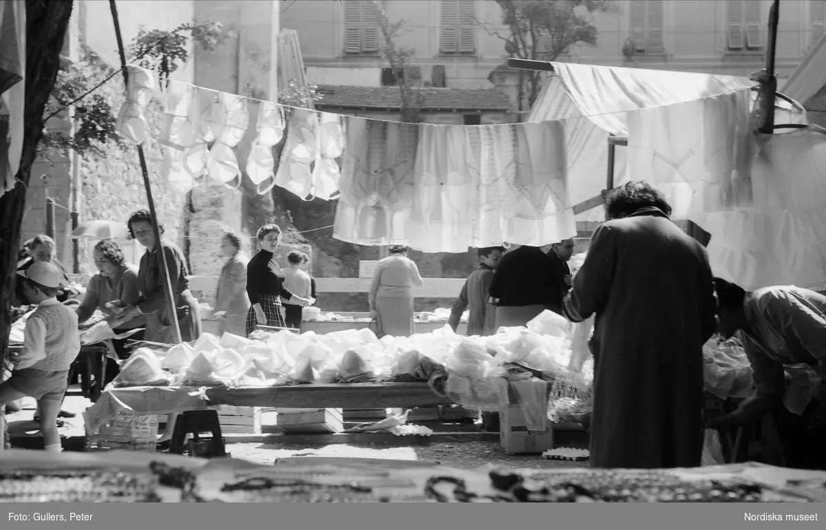 Försäljning av underkläder. BH och underklänningar hänger på lina och ligger på bord. Skärpan ligger på en kvinna en bit bort som vänt sig åt sidan och ser in i kameran. Levanto, Italien