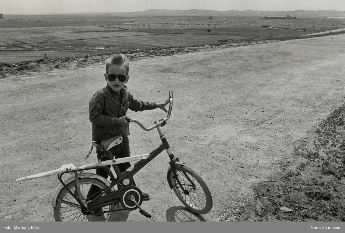 Pojke med randig skjorta och solglasögon håller i en cykel av märket "ABC" på sandväg.
