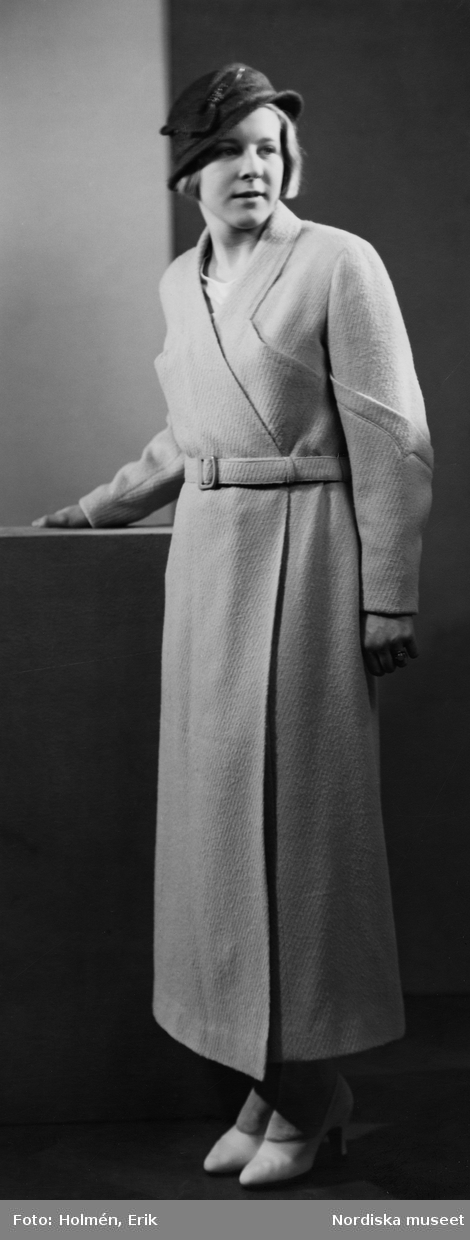 Modell klädd i kappa med skärp, pumps och hatt.