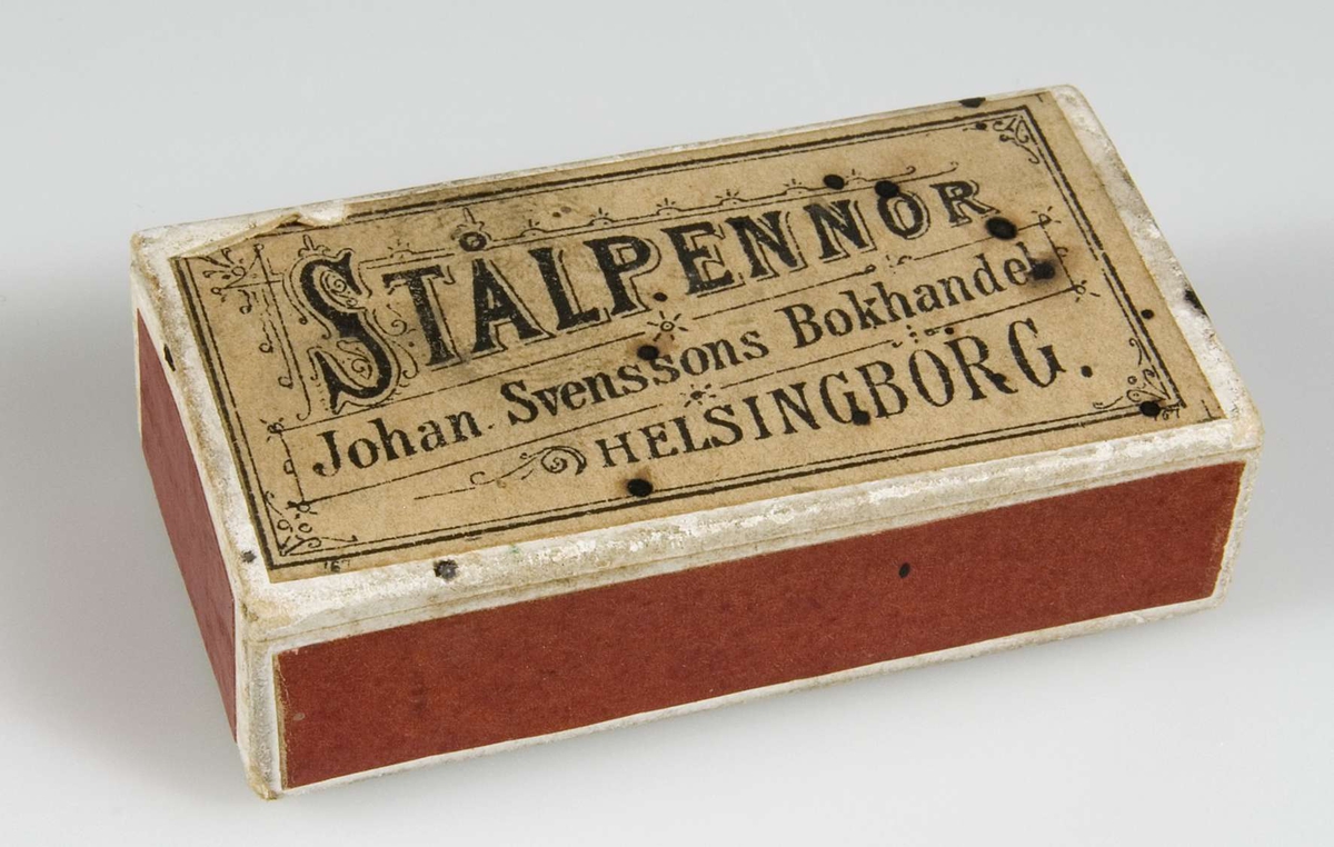 Ask av papp, vit med röda sidor. Vit etikett med svart text: Stålpennor, Johan Svenssons Bokhandel, Helsingborg.