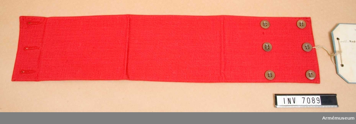 Sydd i rött bomullstyg med reglerbar knäppning under ärmen i form av tre rader knappar. Bindeln är fodrad med ett  bomullstyg i något gulare nyans än yttersidans röda ton. Vidhängande lapp med text: Arbetsmodell å armbindel m/42 för stationsoffgrp (röd).