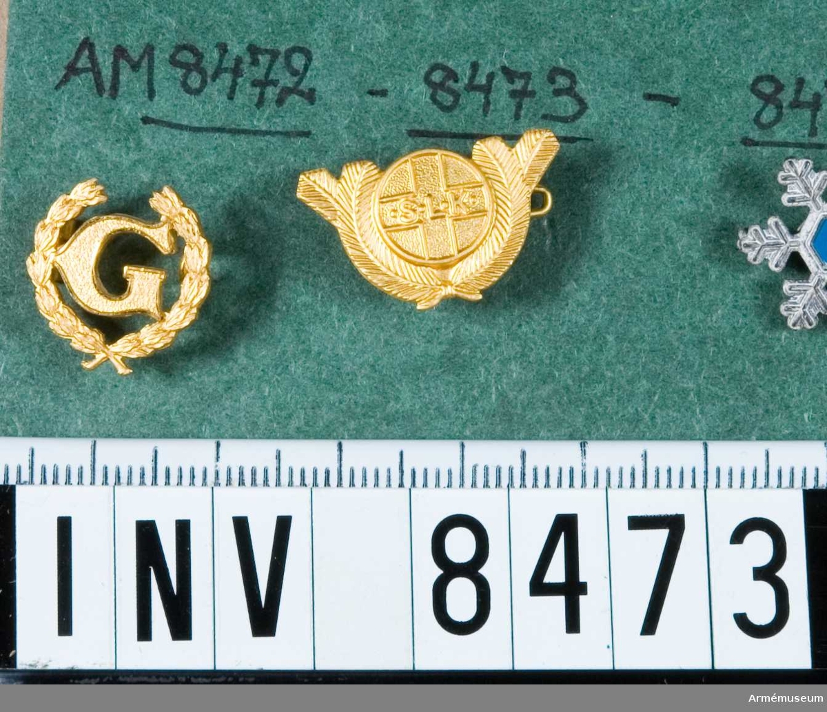 Samhörande nummer är 8469-8474.
Orienringsmärke i guld, d v s förgyllt. För unglottor inom SLK. Märket erövrat 1960. Märket är så gott som runt och platt. I mitten står bokstäverna S.L.K. Bokstäverna är omgivna av en kvist, förmodligen gran.