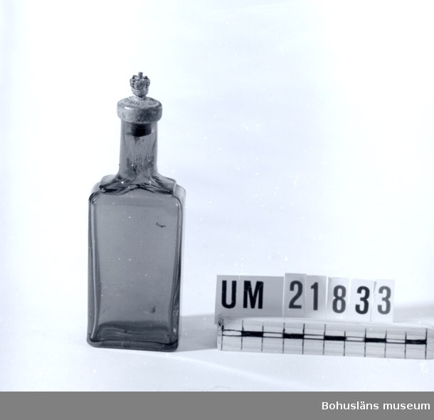 Flaskan rektangulär med rund hals och droppkork av vit metall. Blåfärgat glas.