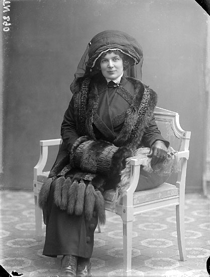 Enligt senare noteringar: "Ateljéfoto av kvinna iklädd dräkt med stor hatt med flor, boa och muff av pälsverk."