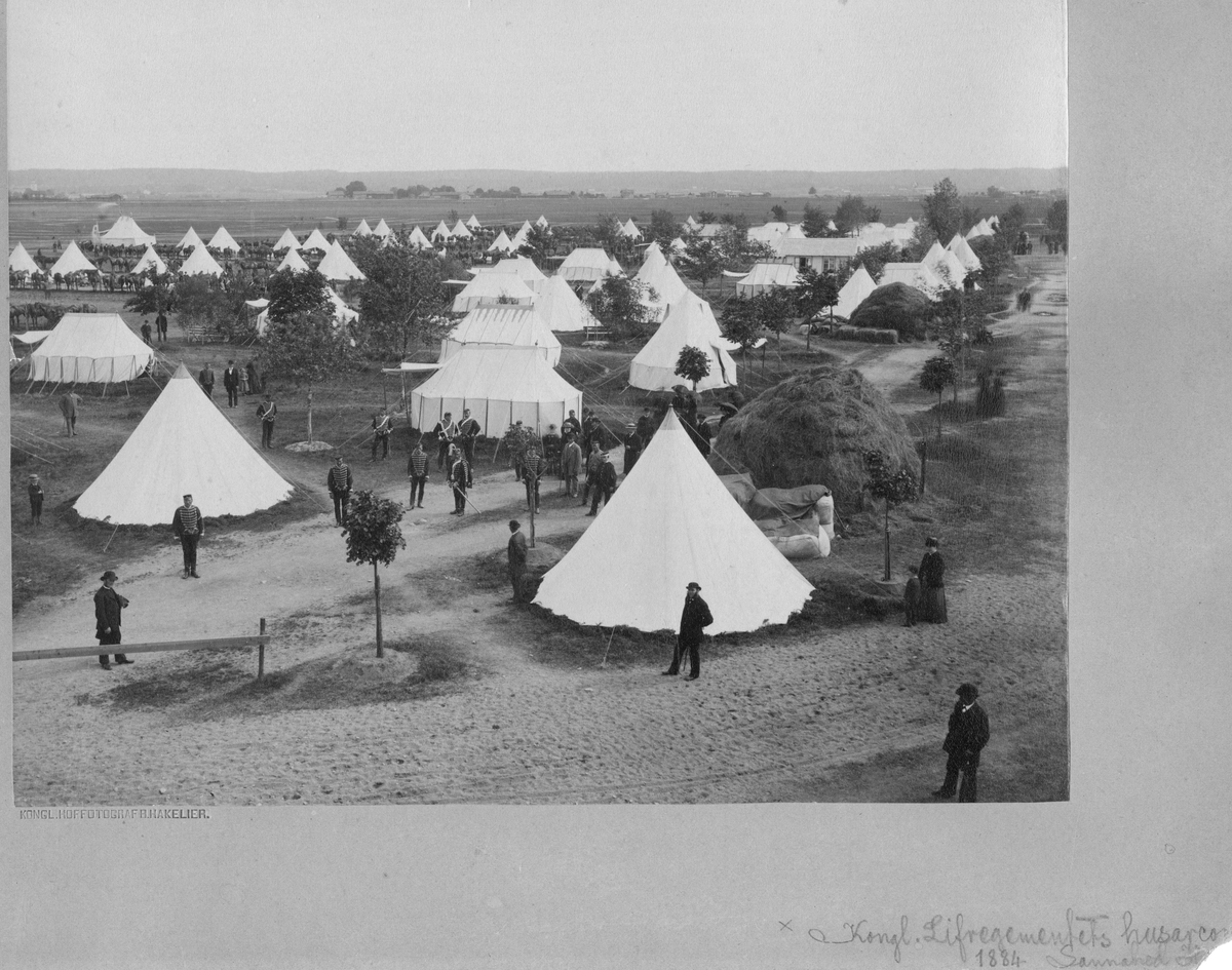 Soldater från Livregementets husarer K 3 står utanför tältläger på Sanna hed. Utanför några tält ligger stora höbalar. Civila personer är på besök i lägret.