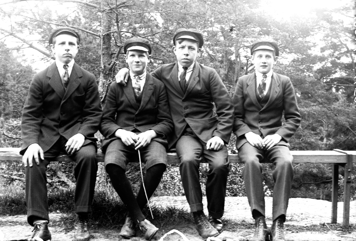 Fyra pojkar sitter på en bänk. De är klädda i kostym och slips och med skärmmössa på huvudet. Pojke nr två håller en pinne i handen. I bakgrunden växer träd och buskar.