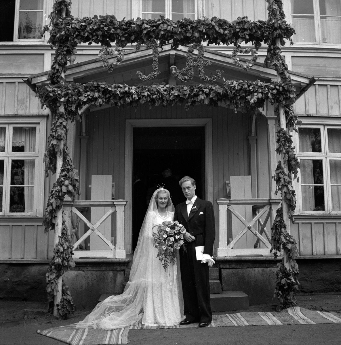 Bröllop på Granbäcks gård mellan Ingrid Qvänstedt och Arne Dalefält i juni 1948. Hon i lång spetsklänning med rosor och prästkragar i buketten. De står under äreporten av gröna bladgirlander med initialerna I och H.