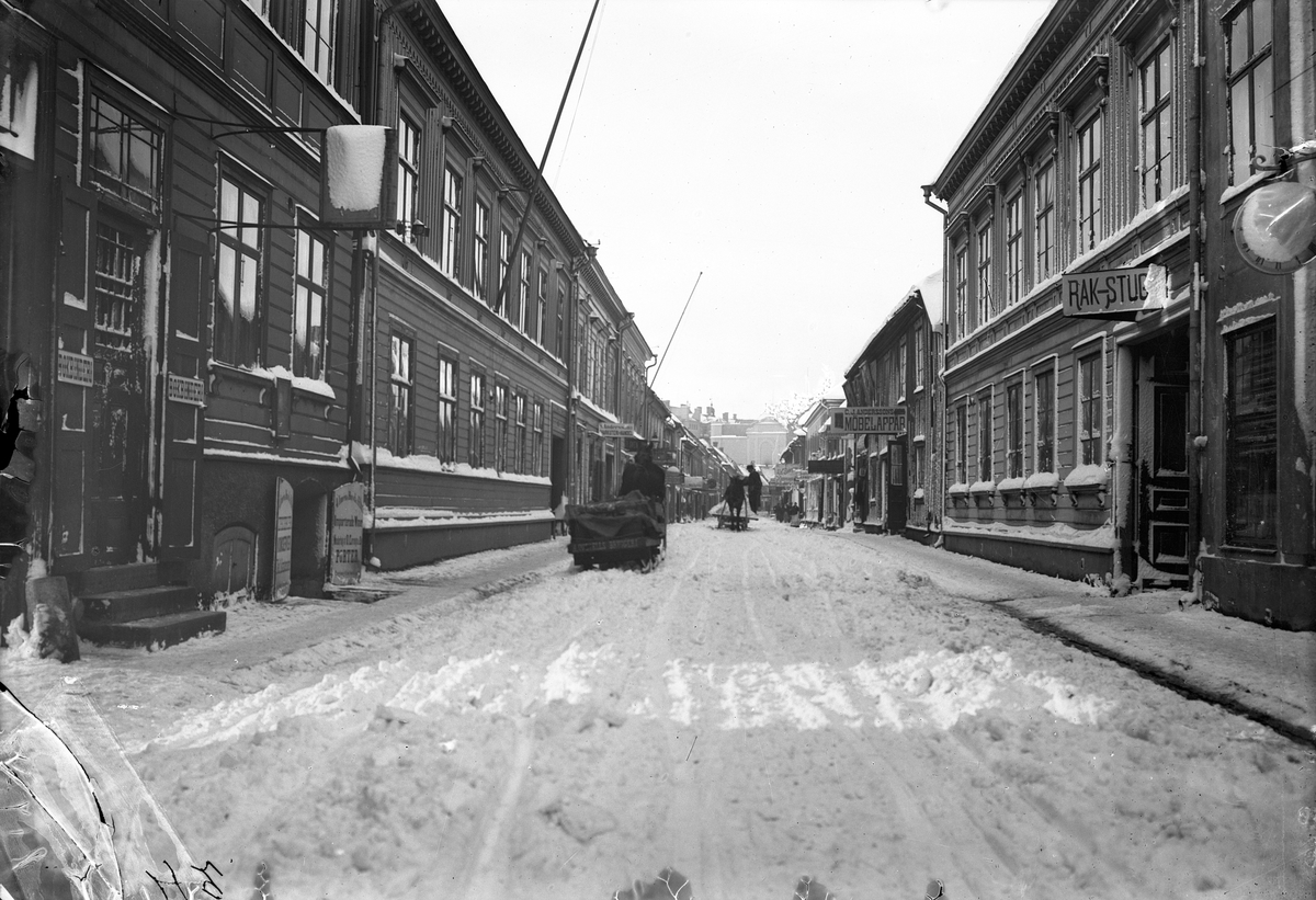 Smedjegatan västerut från Hovrättstorget i Jönköping omkring år 1900. Till höger ligger en rakstuga och längre bort på gatan C.J.Anderssons möbelaffär. Till vänster finns ett bokbinderi och i källaren bredvid kan man köpa sig en porter med mera.