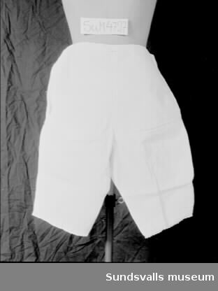 Benkläder i vitt bomullstyg. Sprund i var sida som knäpps med en knapp. Nederst på benen två lagda veck, samt brodyr med hålsöm och uddkant.