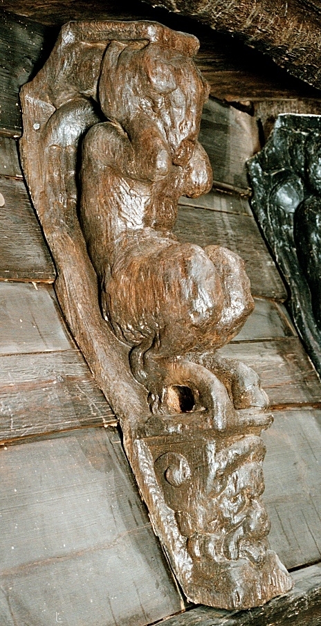 Skulptur föreställande en djävulsfigur i starkt hukande ställning, placerad på en hög sockel.
Figurens huvud är utrustat med stora, krumma horn. I vardera handen håller den på ömse sidor av huvudet något som möjligen kan tolkas som en stor snäcka eller lur. Hakan pryds av ett bockskägg. Figuren har fladdermusvingar på ryggen och starkt luden underkropp med klövförsedda bockfötter. Fötterna vilar på den starkt sluttande sockeln, som framtill är prydd med en ap- eller lejonliknande maskaron med stor nos och hög, välvd panna. På sockelns sidor ses voluter.
Sockeln utgör fortsättning på det kraftiga klink-konstruerade bakstycket som är framdraget ovanför figuren.
Skulpturen är delvis kraftigt sliten och vittrad, särskilt den övre delen. Nederdelen är något bättre bevarad.

Text in English: Sculpture of a devil figure in a crouched position, placed on a high socle.
The figure''s head is equipped with large, curved horns. In either hand the figure holds on either side of its head something that may possibly be identified as a large seashell or horn. The devil has a goat''s beard on its chin, bat wings on its back and very hirsute lower parts of the body, and cloven goats'' feet.
The feet rests on a sharply sloping socle that is decked in front with a mask of an ape-like or lion-like face with a large nose and a high arched forehead. On the sides of the socle volutes are visible.
The socle forms the extension of the heavy, clinker-built back-piece, which is drawn forward above the figure.
The sculpture, particularly its upper parts, is much worn and disintegrated. The lower part is somewhat better preserved.