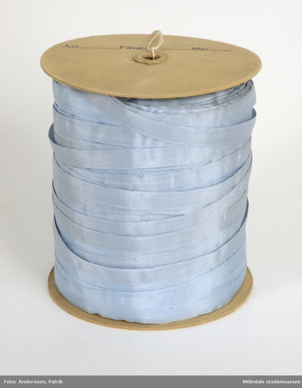 Ljusblått sidenband på rulle, som använts vid tillverkningen av produkter på Kuverteringen, Papyrus. Bandet är lindat på en papprulle med rund platta i var ända. Ena sidan är märkt med blyerts "Ti (texten är något otydlig) 1242/24, blå cp., 500". "Blå", avser färgen på bandet och "500" är bandets längd på rullen i meter. 