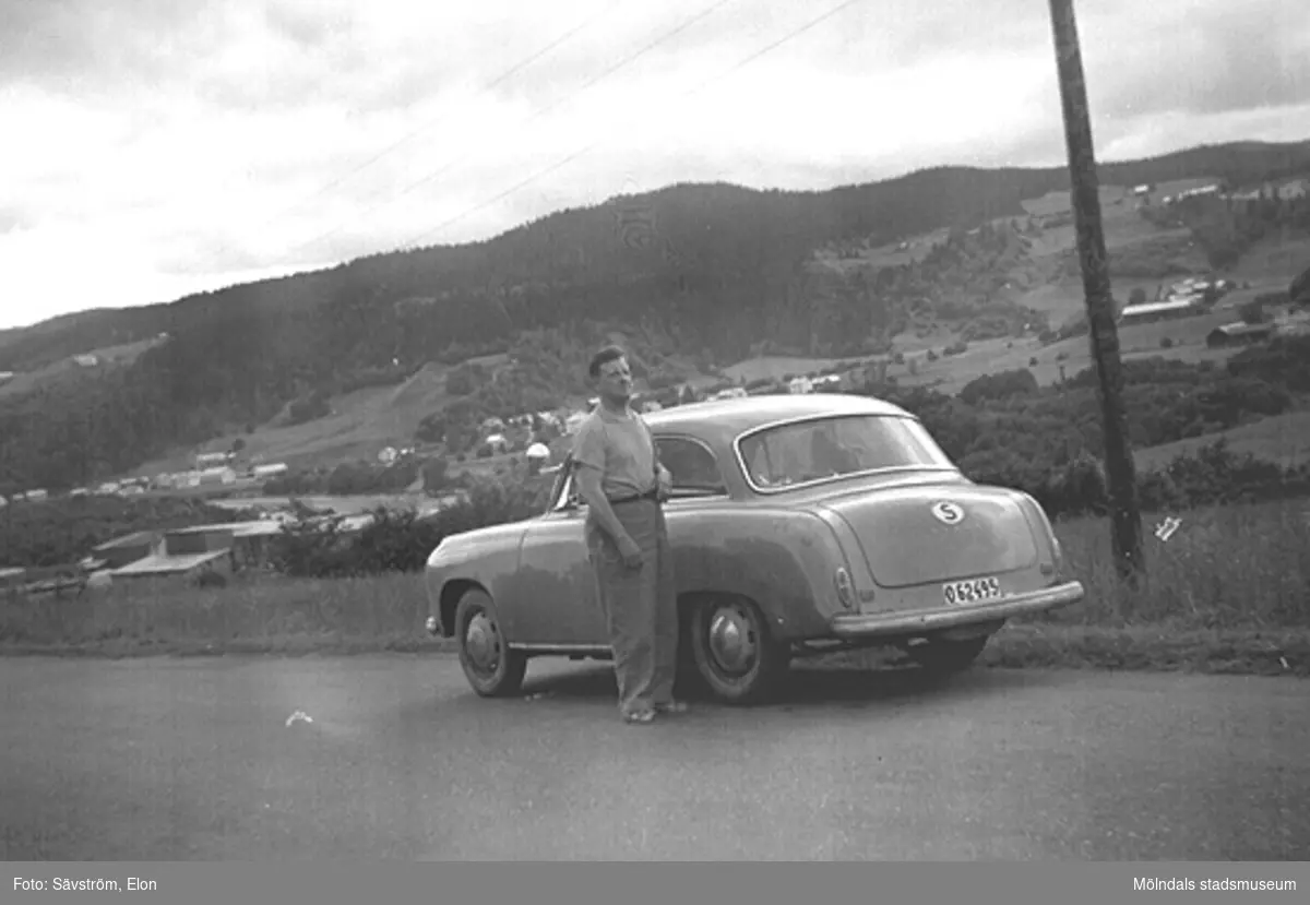 Helmer Garthman är på väg till Norge för fiskesemester med Elon Sävström och dennes bil, en Goliat Isabella. Här är det paus i Värmland. 1960-tal. Skänkt av sonen Alf Garthman som arbetade många år på Mölndals stadsmuseum.