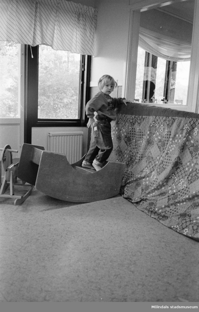 Inomhus på Katrinebergs daghem. Ett barn står på en trägunga och lutar sig mot en stående mjuk madrass som är överdragen med ett tyg. I bakgrunden syns en gunghäst samt fönster med gardiner.