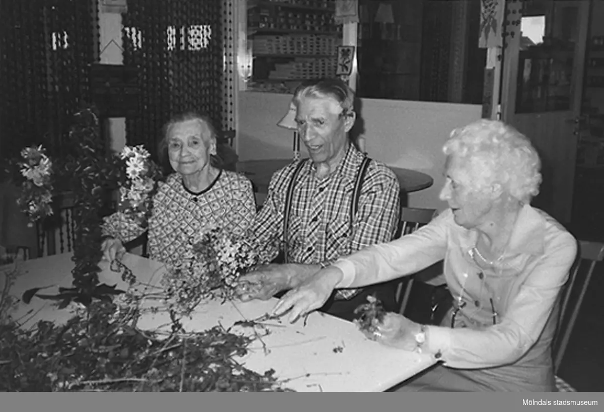 Två kvinnor och en man plockar bland blommor. De klär en liten midsommarstång som står på bordet framför dem. Okänt årtal.