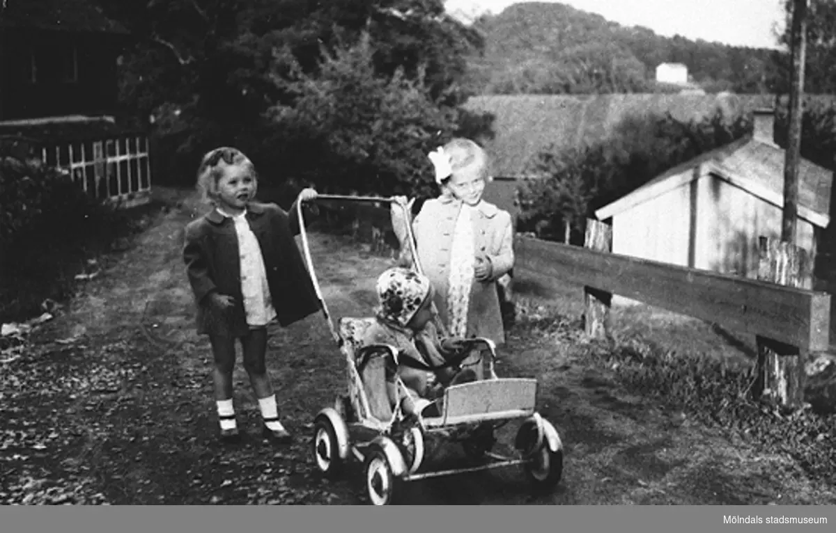 Tre vänner på promenad vid Torrekulla gård, midsommar 1948. Från vänster: Gerd (vän på besök), Eva Pettersson (gift Kempe) och lillasyster Karin Pettersson (gift Hansson) som sitter i kärran. I bakgrunden syns lillstugan samt Brattåsberget vid Kållereds centrum.