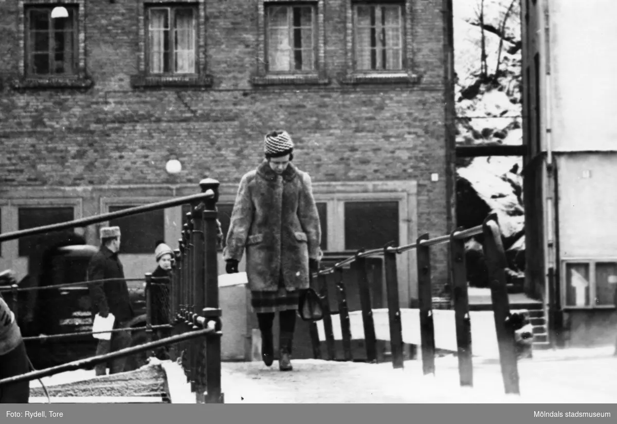 En kvinna promenerar över Forsebron i Mölndals Kvarnby en vinterdag ca 1966-1967. I bakgrunden ses byggnaden Kvarnbygatan 41, tidigare brandstation och garage för polisbilar, numera Mölndals målarskola.