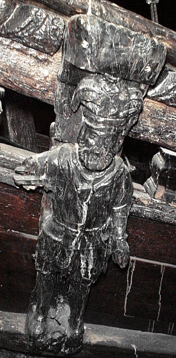 Skulptur föreställande en mansfigur i rustning. På huvudet har figuren en plymförsedd hjälm med taggig krona. Ansiktet är fyrkantigt med långa mustascher och kort, tvärklippt skägg. Figuren är iförd en renässansrustning med hamrat böstharnesk, lårskört och knäskydd. Manteln hänger ned utefter ryggen och är ihopfäst runt halsen med ett stort spänne.
Figuren är frontalställd med huvudet vridet en aning mot höger. Kroppstyngden vilar på båda benen. Höger arm saknas, men har hållts lyft utmed sidan. Högerhanden bör ha gripit om en lång lans, som dock saknas. Vänster arm hålls något böjd, med handen placerad vid höften. Ett litet fågelhuvud vid handen utgör rest av ett svärdsfäste. Figuren står på en sockel, vilken pryds av en grotesk maskaron samt en namnbanderoll.
Upptill är bakstycket framdraget i en stor "huvudplatta", försedd med en kraftigt förtjockad och odekorerad framkant. Huvudplattans profil är delvis kvartscirkelformad.
Skulpturen är relativt välbevarad, men kraftigt ytsliten. Sockeln är skadad och namninskriptionen är oläsbar. Figurens fötter är slitna. Höger arm saknas.

Text in English: A sculpture representing a male figure in armour. On the head there is a helmet with plumes and a crown with points. The square-shaped face has long mustaches and a short clipped beard. The figure is wearing armour of renaissance or early sevententh-century type, with a hammered breast cuirass and thigh and knee guards. The mantle hangs down the back and is fastened around the neck with a large buckle.



The figure faces front with the head turned slightly to the right. The weight of the body is evenly distributed over both legs. The right arm is now missing but, at the time the sculpture was salvaged, the right arm was raised to the side and the hand appeared to have once held a lance. The lance is also missing. The left arm is bent with the hand at hip level holding what appears to be the remains of a scabbard decorated with a birds head.The figure stands on a plinth which is decorated with a grotesque mask and a label.



The board at the back extends obliquely upwards in a large and plain "head board".

The sculpture is rather well preserved but very surface worn. The plinth is badly damaged and the letters on the label are not visible. The figure''s feet are worn, and its right arm is missing.