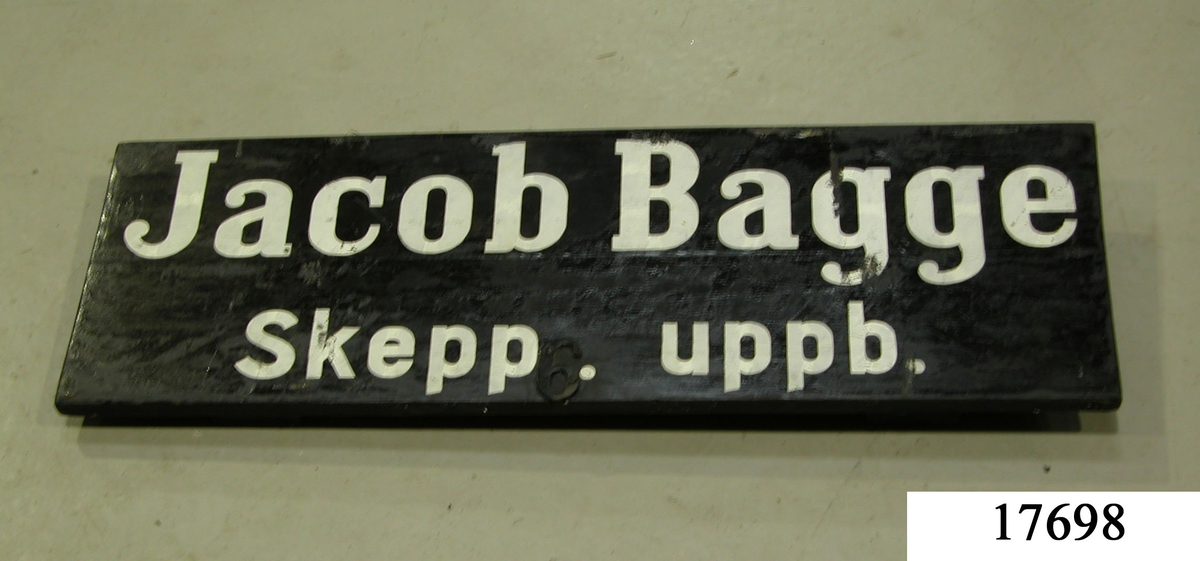 Skylt av träplankor, sammanfästa med brädor på baksidan. Svartmålade, vit schablonmålad text: " Jacob Bagge skepp. uppb.". I skyltens mitt sitter siffran 6 fastskruvad.
