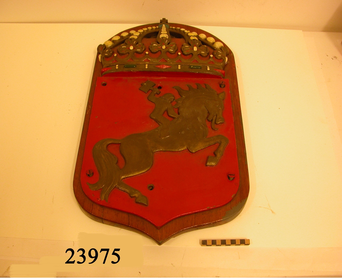 Vapensköld gjuten av mässing, krönt med kunglig krona, målad i heraldiska färger. Botten i rött, där på en galopperande guldförgylld häst med ryttare som höjer en hammare. Skölden är fästad vid fernissad platta av trä. På plattans baksida urfrästa fördjupningar för att passa mot skrovets nitar vid upphängning samt skrivet med tusch: SSS 266.