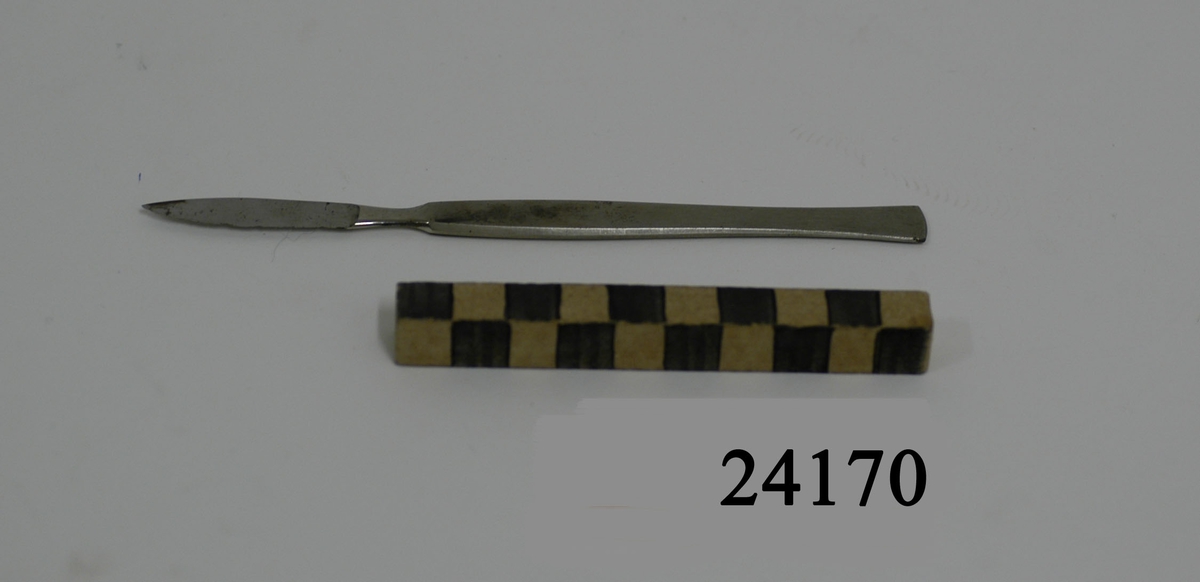Tunn platt operationskniv. Skaft av borstat stål, något insvängt i den nedre delen av skaftet. Knivbladet blankt, eneggat.