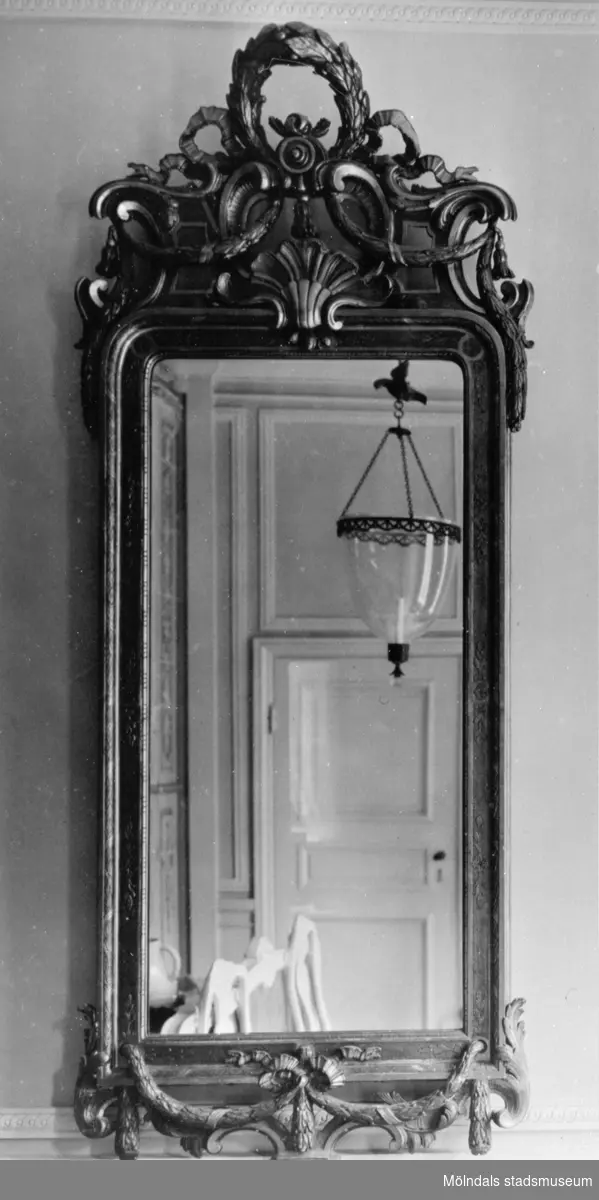 Vägghängd avlång spegel med dekor. I spegelglaset ses delar av ett rum med enkeldörr och taklampa. Gunnebo slott 1930-tal.
