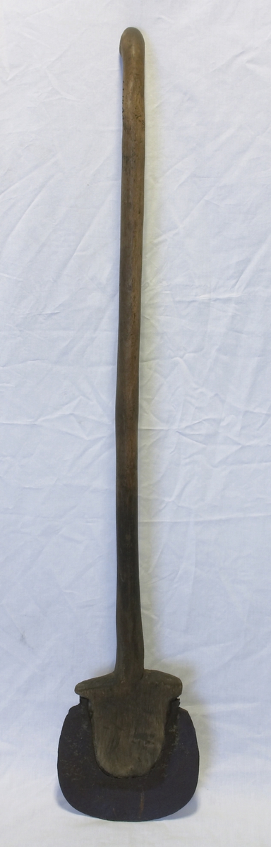 Et langt runlig skaft av tre som øverst bøyer bakover til et håndtak, nederst går det ut i et treblad (spade) som det er festet en jernbue rundt. 