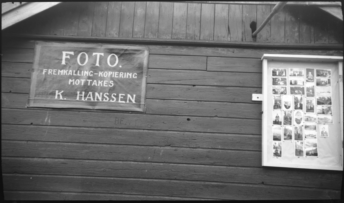 Husvegg med reklameskilt for fotograf Karl Hansen, og eksempler på produktene hans til høyre på veggen.