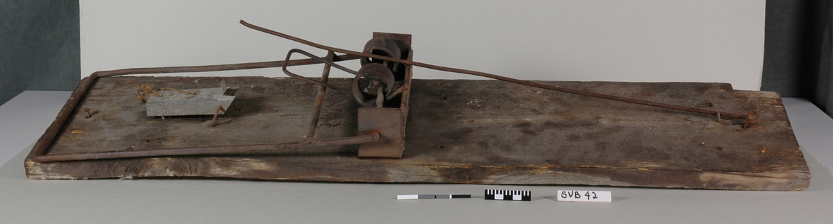 Laget av jern, montert på treplate, umalt. Rottefelleprinsippet. Mekanismen er noe rustet, men ser ut til å kunne fungere.