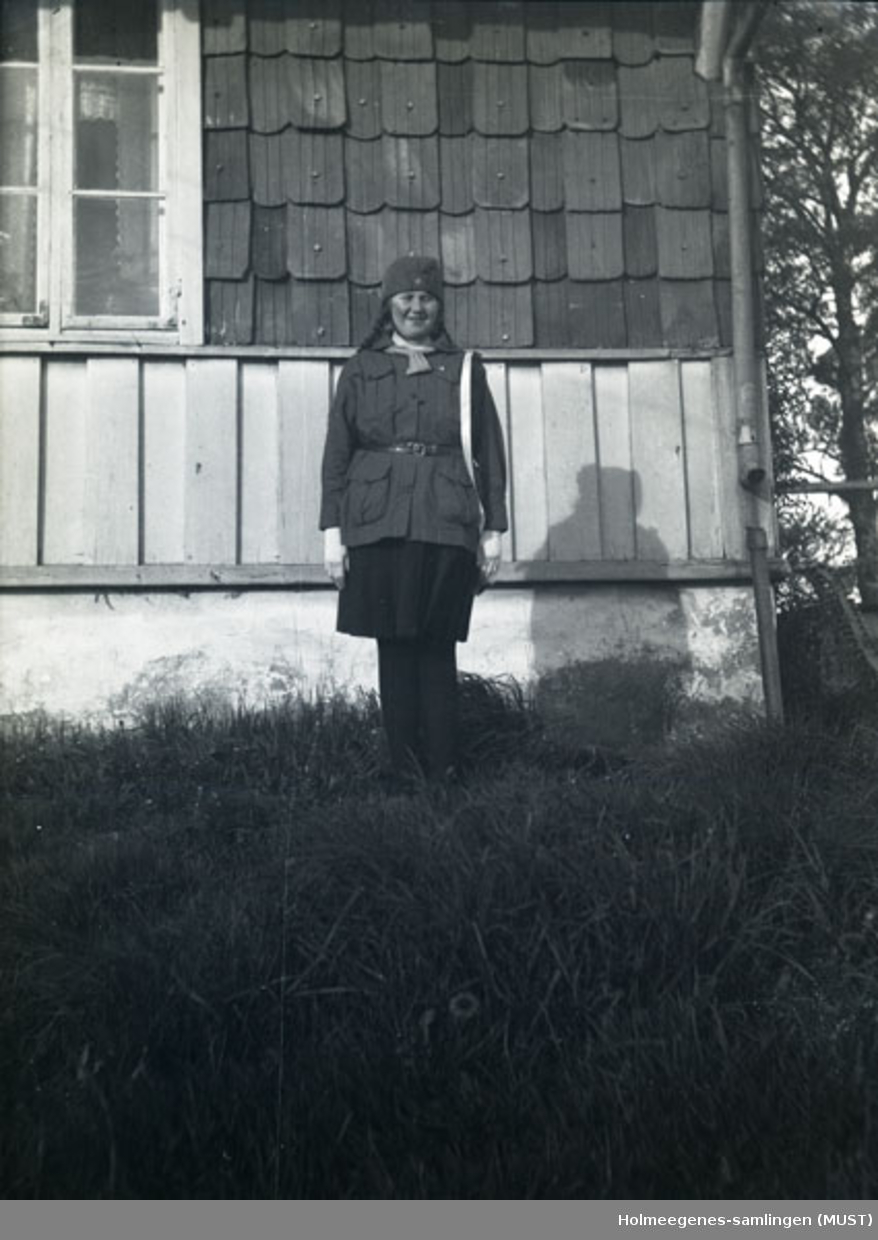 Gunhild Pedersen i speideruniform, står ved et hjørne av huset på Holmeegenes.