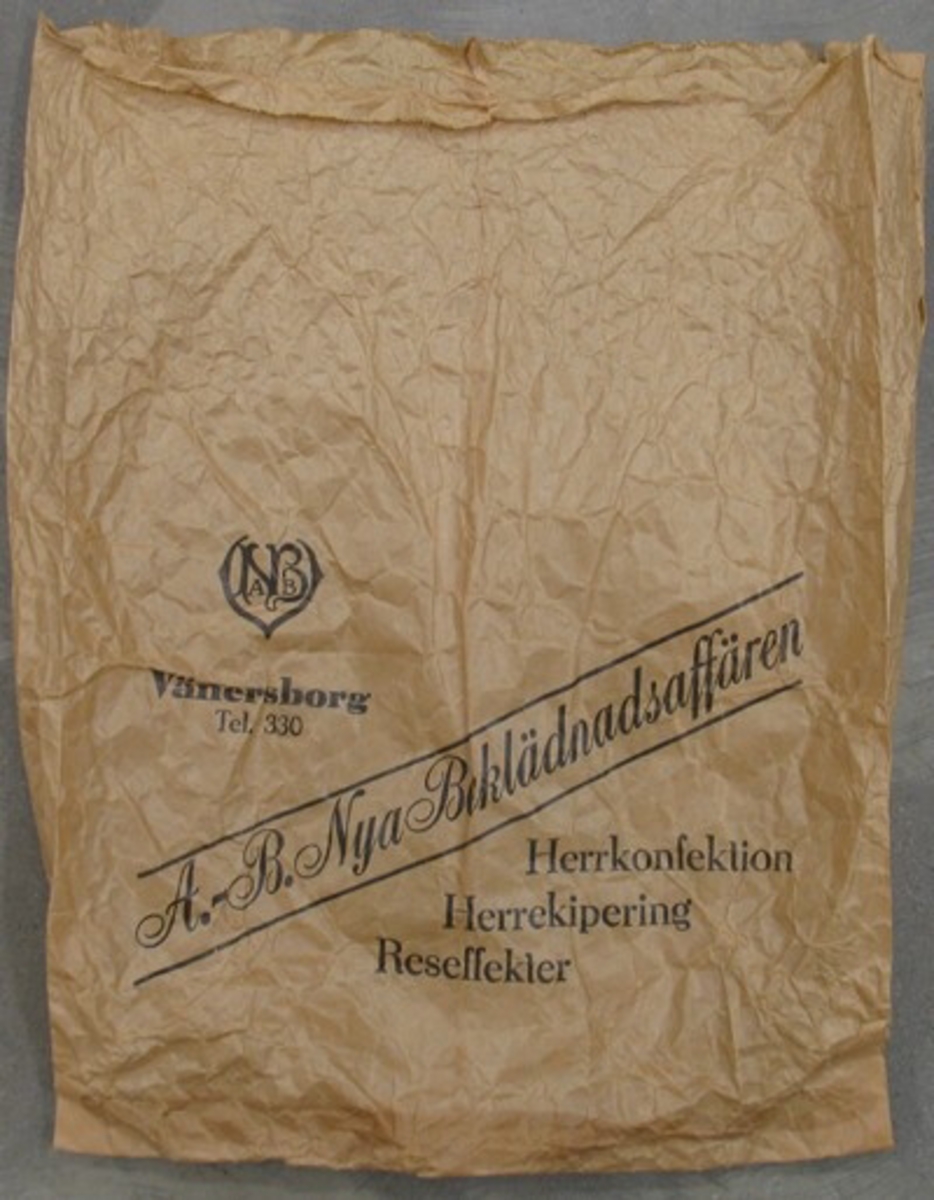 Påse av grått papper med texten: ''A-B Nya Beklädnadsaffären. Vänersborg Tel. 330 Herrkonfektion, Herrekipering, Reseffekter.'' Påsen medföljde inköpet av damhatten VM 21 760.
