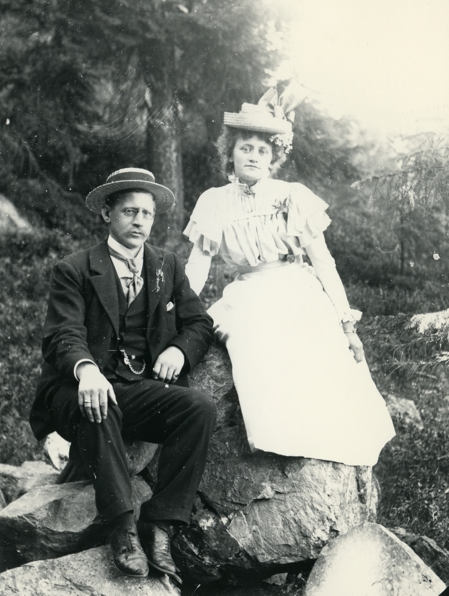 Mann i dress og hatt, og kvine kledd i lys kjole og hatt, med blomster i håret, fotografert på steiner i skog