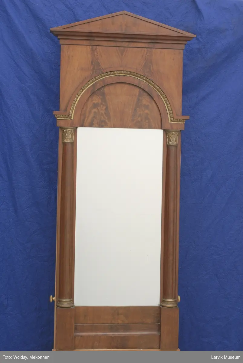 Rektangulært speil i ramme med høyt toppstykke. Toppstykke  med uttrappet røstformet topp. Over speil, rundbue med forgylt list i stukk, båret av to glatte søyler.
