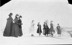 Tre pent kledte kvinner ser på seks jenter som går på ski.
