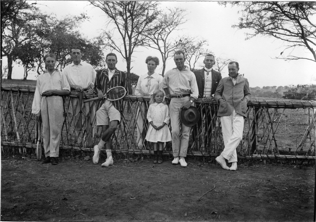 'Diverse fotografier från bl.a. dåvarande Nordrhodesia, nu Zambia, tagna av Konsul Magnus Leijer. ::  :: En kvinna, en flicka och sex män ståendes vid ett staket, tre av männen med tennisracketar. I bakgrunden är höga träd synliga.'