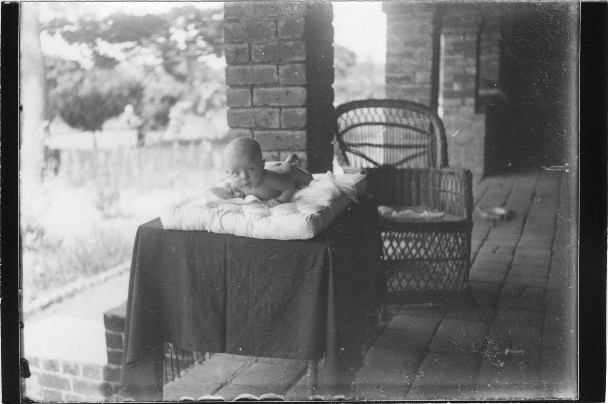 'Diverse fotografier från bl.a. dåvarande Nordrhodesia, nu Zambia, tagna av Konsul Magnus Leijer. ::  :: En naken bebis liggande på en madrass på ett bord, placerat på en stenlagd veranda. Bakom bordet står en rottingfåtölj. Bebisen ser in i kameran.'