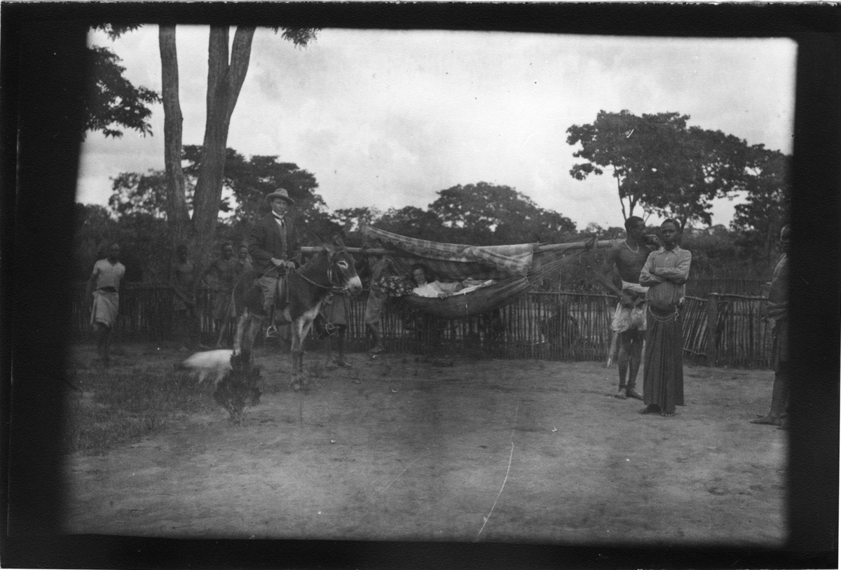 'Diverse fotografier från bl.a. dåvarande Nordrhodesia, nu Zambia, tagna av Konsul Magnus Leijer. ::  :: 2 st män bärande 1 st kvinna i en hängmatta hängande på en trästam som de bär på sina axlar. Ytterligare 3 st män och 1 st man på åsna, synliga.'