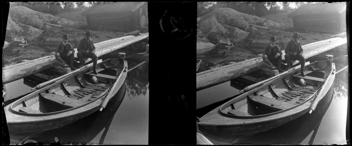 'Bildtext: ''J.E.F.  Och Charleston med 21 gäddor, Sävö.'' :: 2 hattklädda män sittande på brygga, en med fiskespö och den andre med håv. Framför dem 1 båt med åror och motor där 21 gäddor ligger uppradade på bottnen. I bakgrunden skymtar sjöbod. ::  :: Ingår i serie med fotonr. 5255:1-18, se även hela serien med fotonr. 5237-5267.'