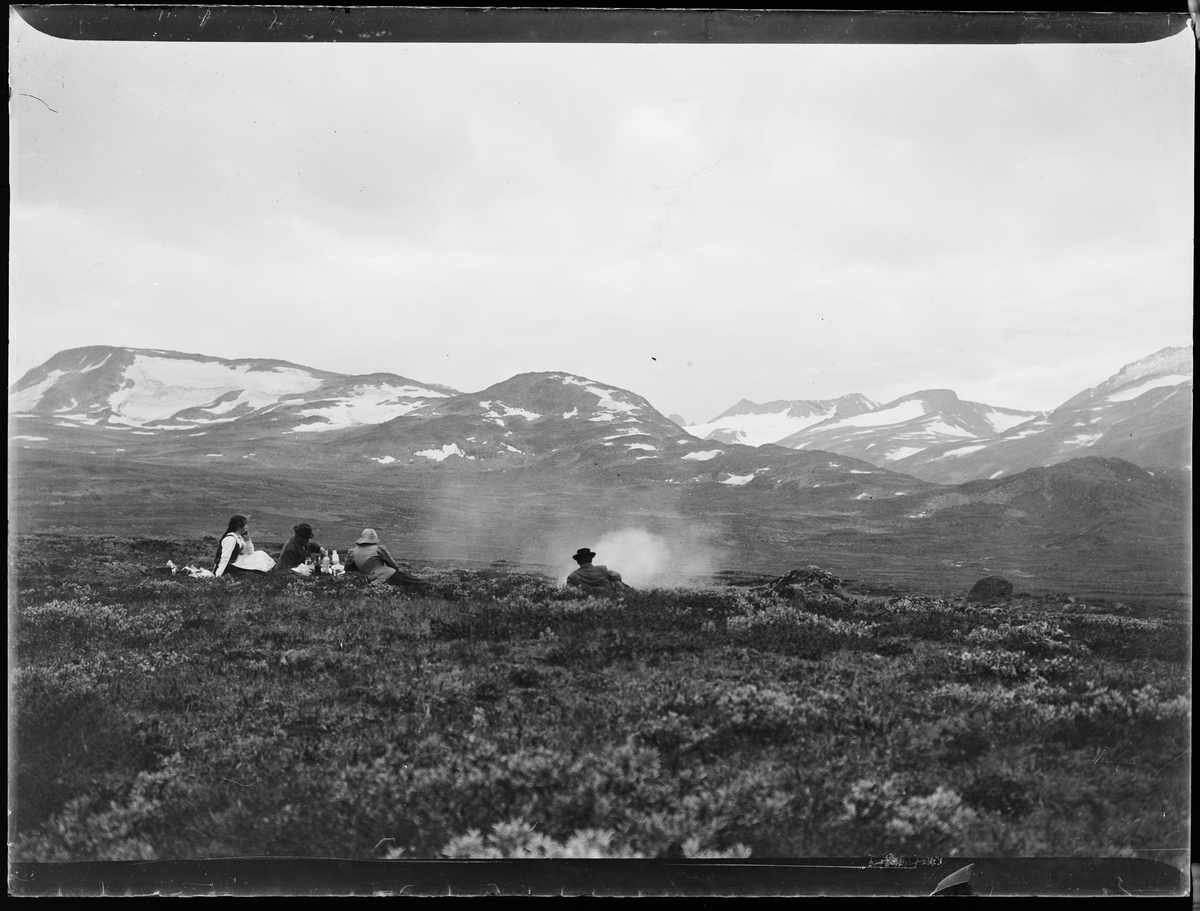"Heimdalen 1906" (inskr eske). Fjell-landskap. Snødekte fjell i bakgrunnen. Fire personer ligger i lyngen. Det ser ut til at de har tent et bål. Alle er vendt fra kameraet.