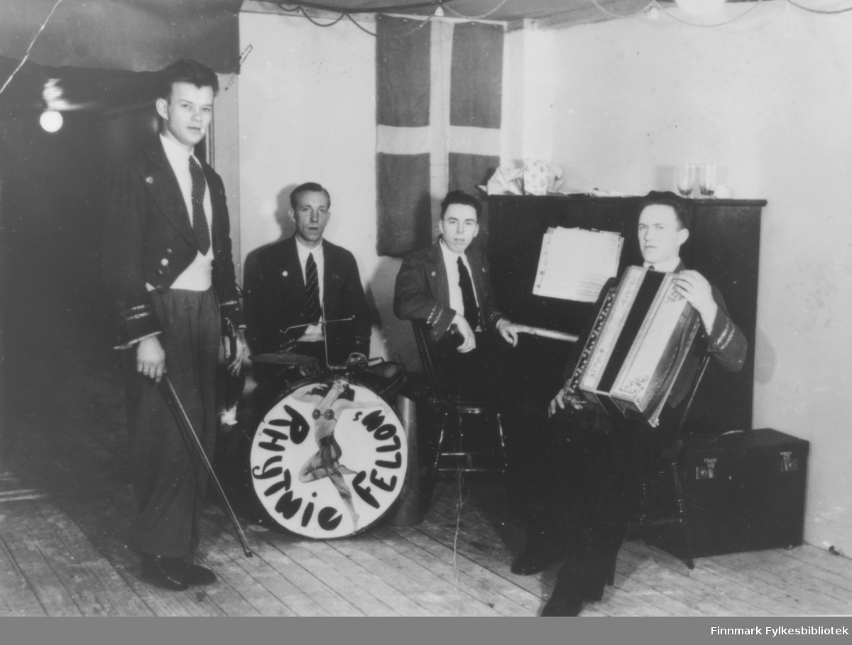 Bilde av 'Rhytmie Fellows' fra Kirkenes. Det står ingen dato på bildet. Fire menn med fiolin, trommer, piano og trekkspill. I bakgrunnen henger et flagg. Det må være dansk, svensk, eller finsk. Gruppen er kledt i dress.