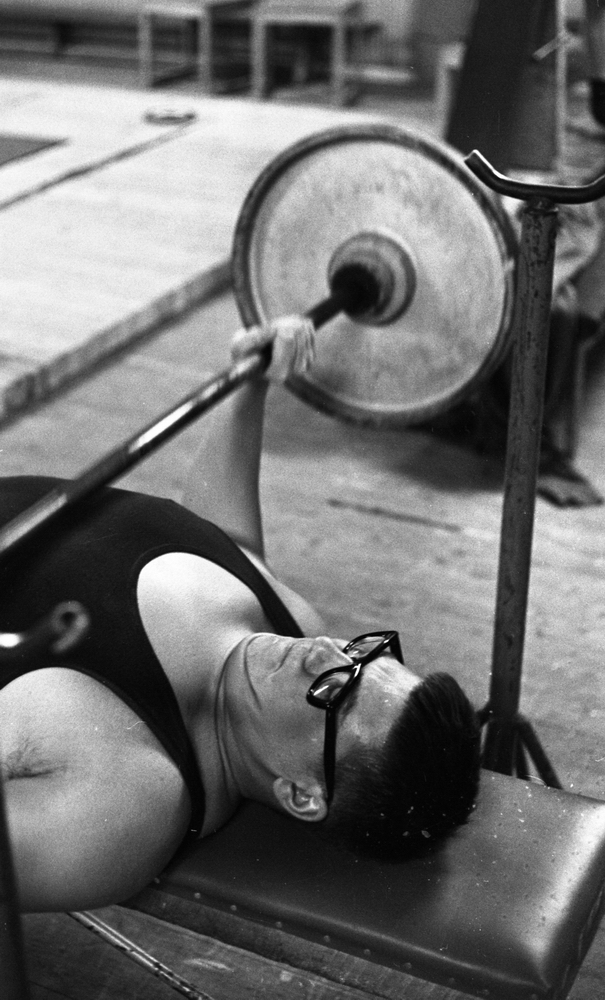 Tyngdlyftarreportage 31 januari 1966 

Närbild på en pojke i äldre tonåren som ligger nära golvet på en bräda och lyfter en skivstång ovanför sitt huvud. Han bär glasögon och är klädd i ett sportlinne.