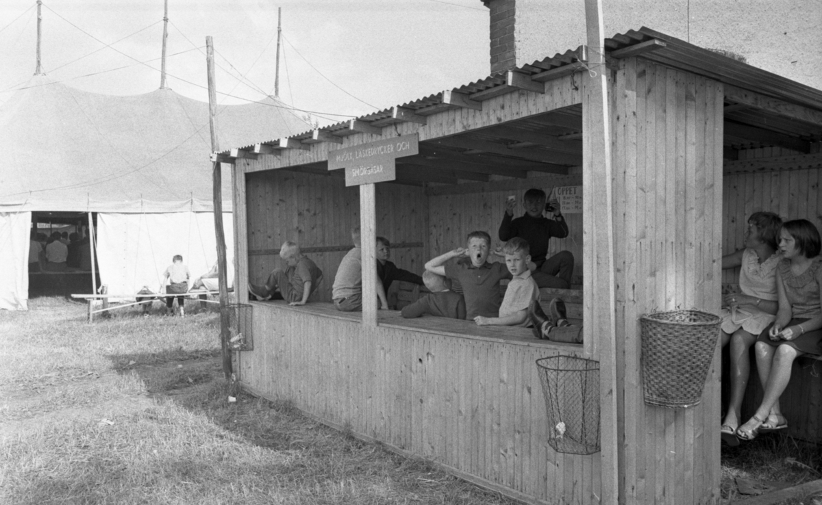 Helgelseförbundet, Japan på Svampen 24 juni 1966

En bild på ett stort, vitt tält. Det är barnens mötestält som tillhör Helgelseförbundet. Genom en tältöppning i bakgrunden syns barn sitta inne i tältet och utanför syns barn sitta på en träbänk. I förgrunden befinner sig några barn inne i en träbod med skylten "Mjölk, läskedrycker och smörgåsar" på. En pojke gör en rolig grimasch mot fotografen.